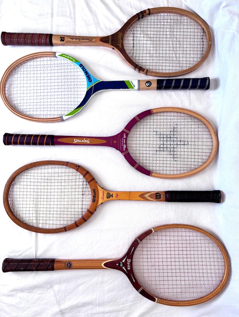 May include: Five vintage wooden tennis rackets with various handle and frame designs. The rackets feature different brand names, including Wilson, Spalding, and Davis. The rackets are arranged on a white surface, showcasing their aged wood and string netting.