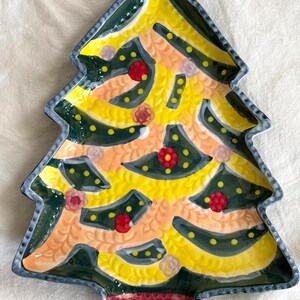 May include: A ceramic Christmas tree-shaped plate with a yellow and green design. The plate features red and pink floral accents and a textured, maroon base. The edges are outlined in a dark blue.