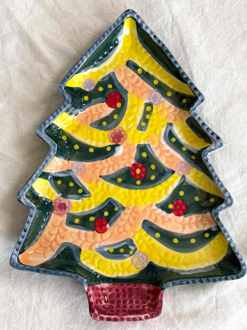 May include: A ceramic Christmas tree-shaped plate with a yellow and green design. The plate features red and pink floral accents and a textured, maroon base. The edges are outlined in a dark blue.