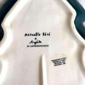 May include: A white ceramic tree-shaped serving dish with a dark green border. The dish features the text "Nathalie L&eacute;t&eacute;" and "Ang&egrave;le for Anthropologie". The dish is hand-painted in China and is microwave and dishwasher safe.