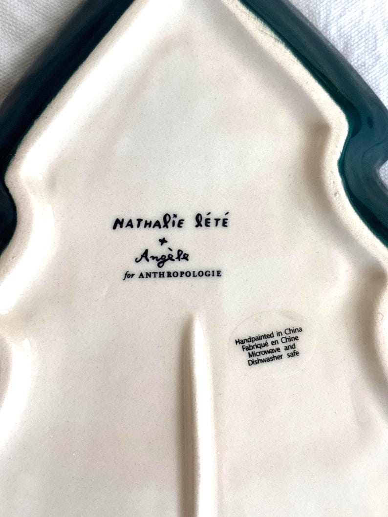 May include: A white ceramic tree-shaped serving dish with a dark green border. The dish features the text "Nathalie L&eacute;t&eacute;" and "Ang&egrave;le for Anthropologie". The dish is hand-painted in China and is microwave and dishwasher safe.