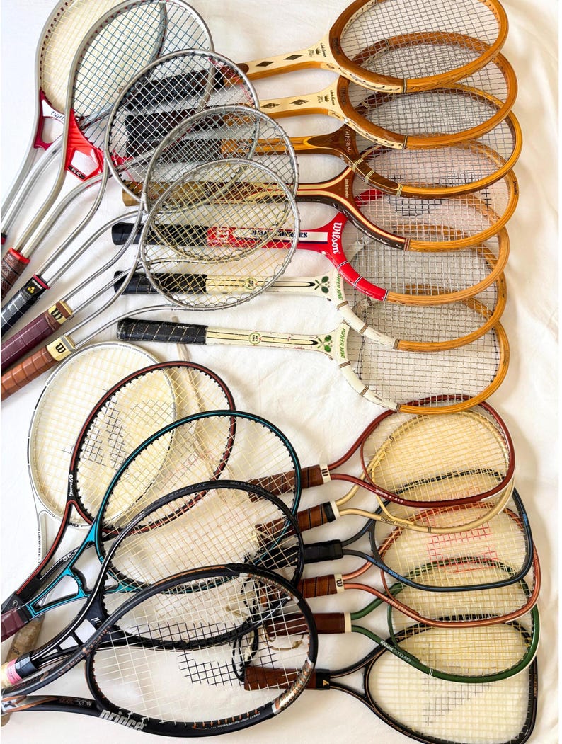 May include: A collection of vintage tennis rackets with wooden and metal frames. The rackets feature various colors, including brown, red, green, and black, with leather grips and strung nets. Some rackets have the "Wilson" brand.