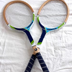 May include: Two vintage tennis rackets crossed, featuring wooden frames and white netting. The rackets have blue and green accents with the word "CHEMOLD" printed on the head. The handles are wrapped in blue grip tape, and the word "Signature" is printed on the side.
