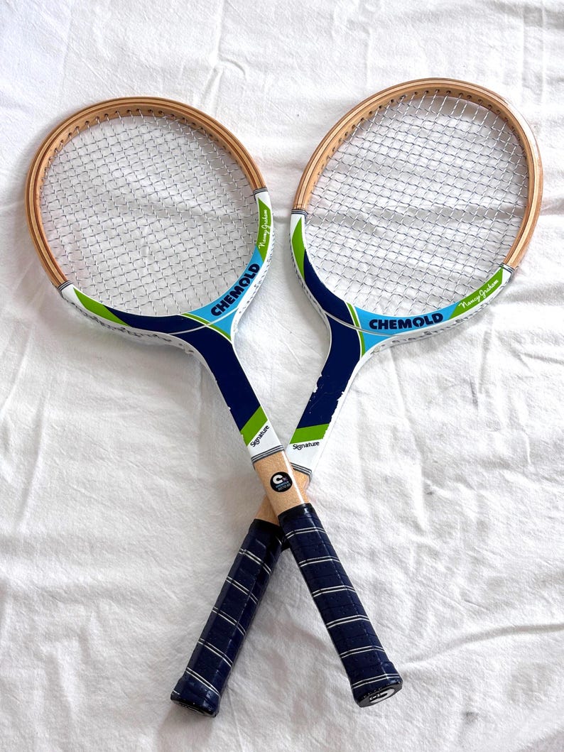 May include: Two vintage tennis rackets crossed, featuring wooden frames and white netting. The rackets have blue and green accents with the word "CHEMOLD" printed on the head. The handles are wrapped in blue grip tape, and the word "Signature" is printed on the side.