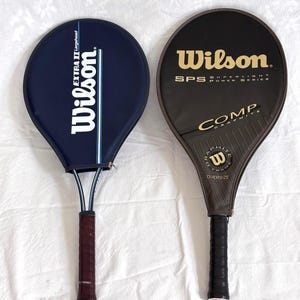 May include: Two vintage Wilson tennis rackets. One is navy blue with white text and a blue stripe. The other is black with gold text. Both have dark brown and black grips.