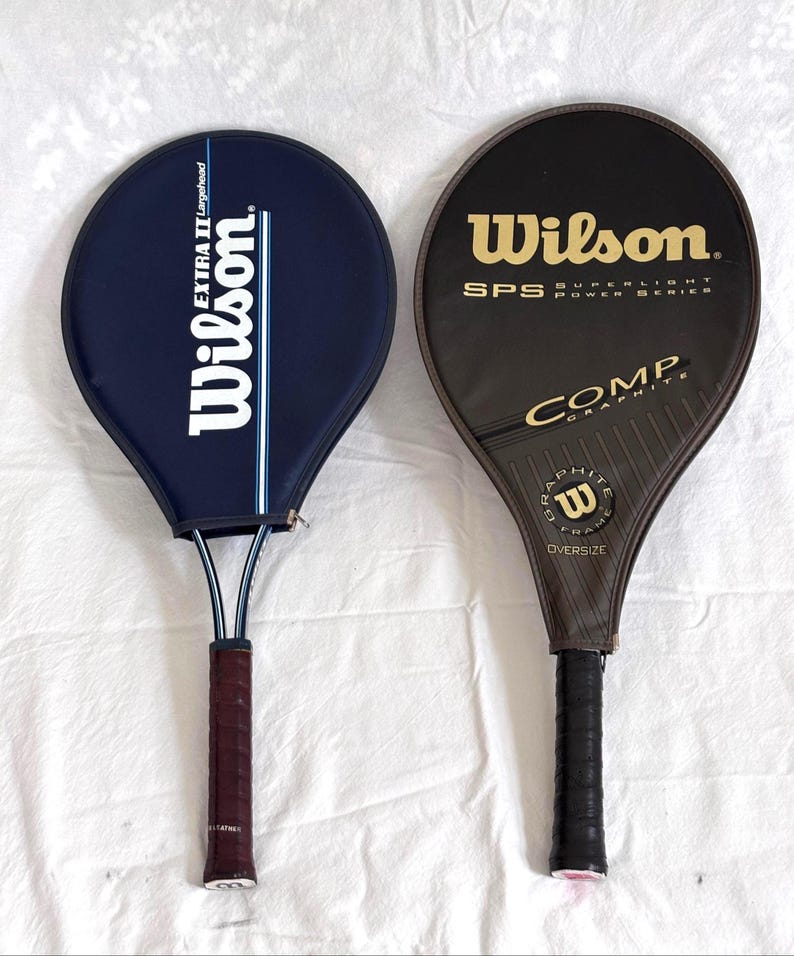 May include: Two vintage Wilson tennis rackets. One is navy blue with white text and a blue stripe. The other is black with gold text. Both have dark brown and black grips.