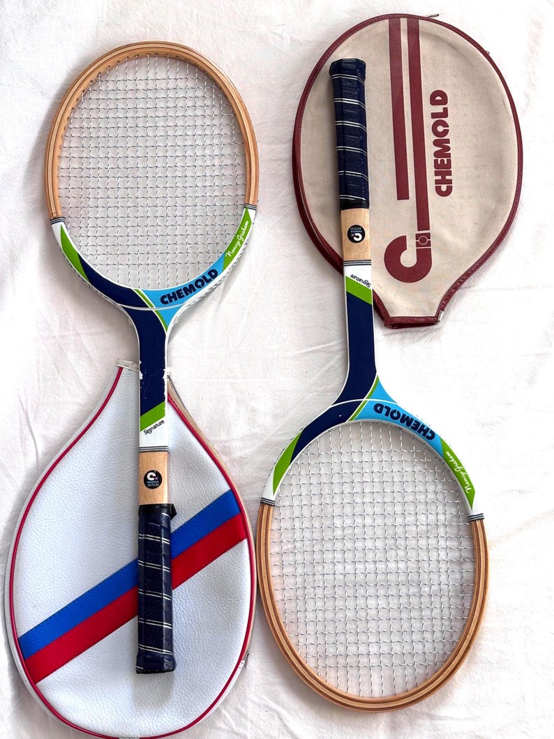 May include: Two vintage Chemold tennis rackets with wooden frames and white mesh. One racket has a blue, green, and white handle with the word "Signature" on it. The other racket has a matching design. Both rackets have matching cases.