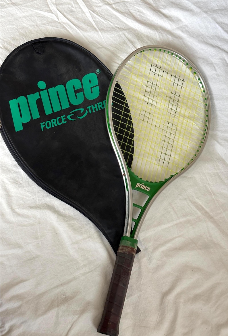 May include: A green and silver Prince tennis racket with a black racket cover. The racket has yellow strings and a brown grip. The racket cover has the word "Prince" in large green letters.