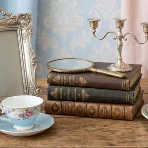 May include: A vintage still life arrangement featuring a silver candelabra, a hand mirror, and a teacup with saucer. The items rest on a stack of antique books. A decorative silver picture frame and a teacup with floral design are also present.