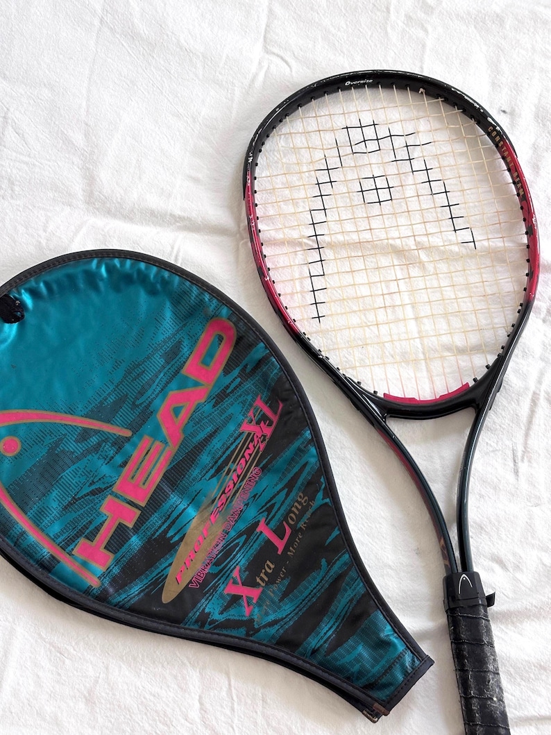 May include: A black and red HEAD tennis racket with a white string net and a black grip. A teal and black racket cover with the HEAD logo and "Xtra Long" text is next to the racket. The racket and cover are on a white surface.