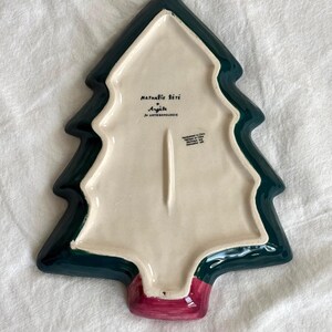 May include: A ceramic Christmas tree-shaped serving dish. The dish features a dark green border, a cream-colored interior, and a red base. The underside has text that reads "Nathalie Lete" and "Ang&egrave;le for Anthropologie."