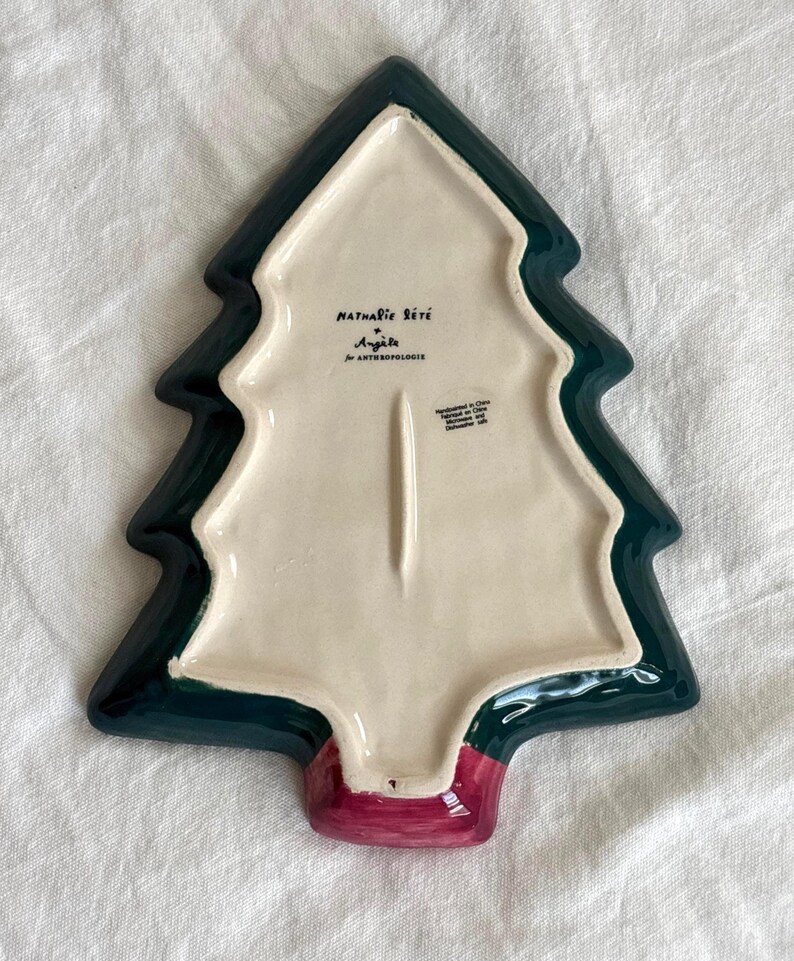 May include: A ceramic Christmas tree-shaped serving dish. The dish features a dark green border, a cream-colored interior, and a red base. The underside has text that reads "Nathalie Lete" and "Ang&egrave;le for Anthropologie."