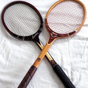 May include: Two vintage tennis rackets with wooden frames and string netting are crossed. One racket is dark brown with "Silverstreak" text, the other is light brown with "Davis" text. Both have leather grips.