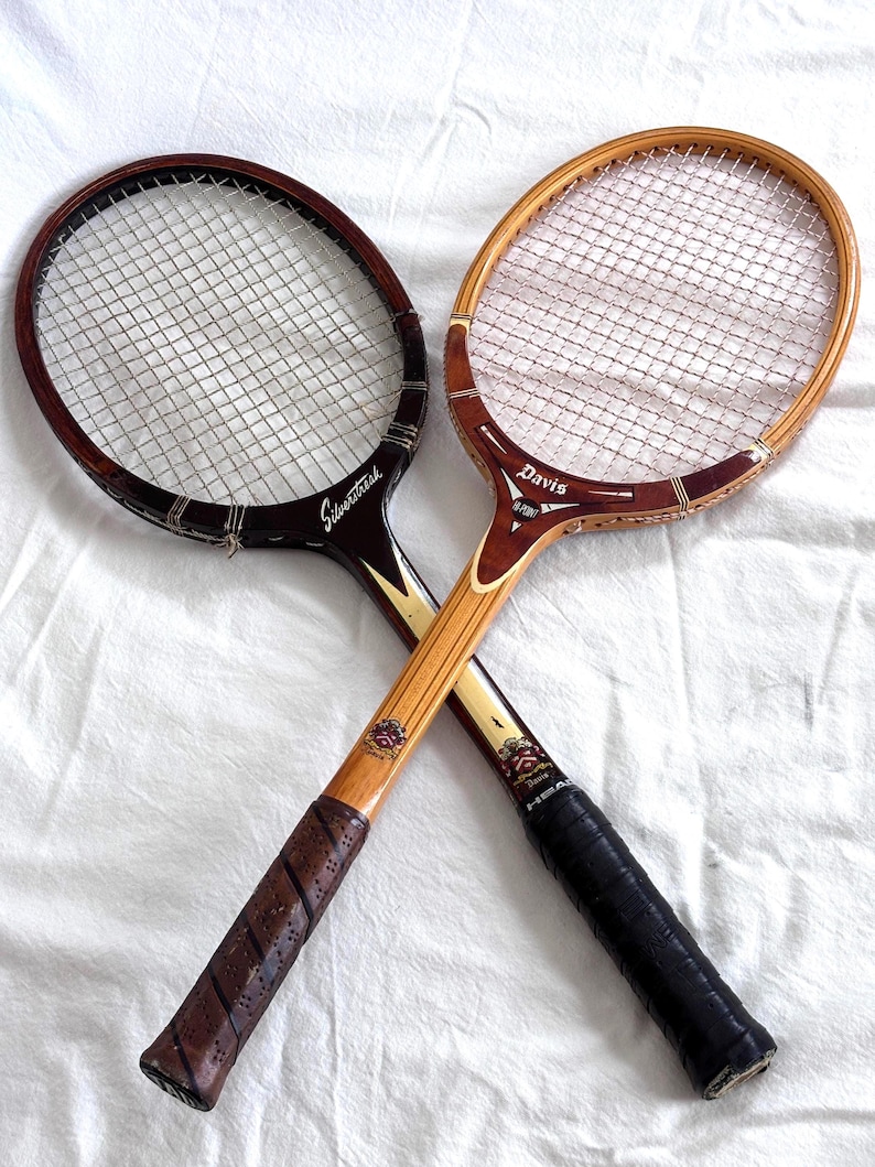May include: Two vintage tennis rackets with wooden frames and string netting are crossed. One racket is dark brown with "Silverstreak" text, the other is light brown with "Davis" text. Both have leather grips.