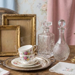 May include: A still life arrangement featuring vintage items. Two gold picture frames, a silver serving tray with floral teacups and saucers, and two crystal decanters. A floral embroidered napkin rests on a wooden surface.