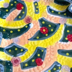 May include: Close-up of a decorative ceramic plate with a vibrant, colorful design. The plate features a yellow background with a textured pattern, accented by dark green shapes with yellow dots, red flower-like details, and light blue edges.