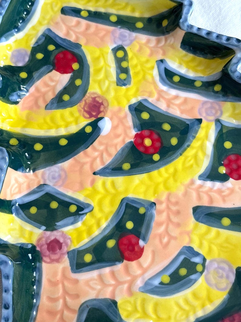 May include: Close-up of a decorative ceramic plate with a vibrant, colorful design. The plate features a yellow background with a textured pattern, accented by dark green shapes with yellow dots, red flower-like details, and light blue edges.