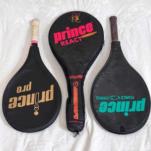 May include: Three vintage black tennis rackets with covers. The rackets have the brand name "Prince" in gold and teal, and the text "React" and "Force Three" are visible. The grips are wrapped in white and pink tape.