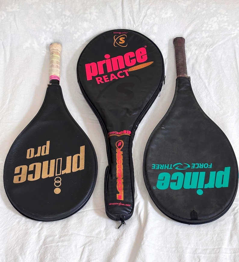 May include: Three vintage black tennis rackets with covers. The rackets have the brand name "Prince" in gold and teal, and the text "React" and "Force Three" are visible. The grips are wrapped in white and pink tape.