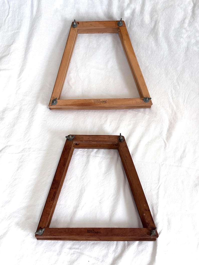 May include: Two vintage wooden tennis racket frames. The frames are trapezoidal, with rounded corners and metal hardware. The frames are different shades of brown, and the word "Wilson" is printed on the bottom of each frame.