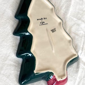 May include: A ceramic Christmas tree-shaped serving dish. The dish is dark green with a cream-colored interior and a red base. The dish has the text "Nathalie Lete" and "Anjole" printed on the interior.