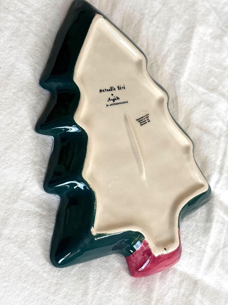 May include: A ceramic Christmas tree-shaped serving dish. The dish is dark green with a cream-colored interior and a red base. The dish has the text "Nathalie Lete" and "Anjole" printed on the interior.