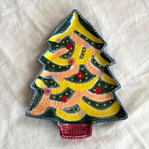 May include: A ceramic Christmas tree-shaped plate with a yellow and green design. The plate features red and pink accents, a dark green border, and a red base. The plate is designed for serving holiday treats.