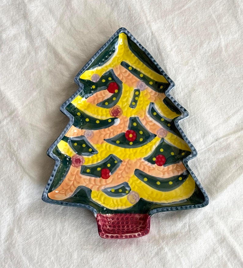 May include: A ceramic Christmas tree-shaped plate with a yellow and green design. The plate features red and pink accents, a dark green border, and a red base. The plate is designed for serving holiday treats.