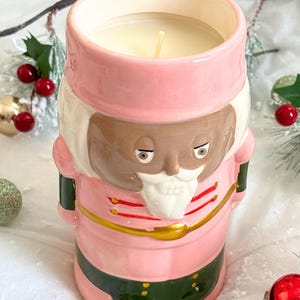 May include: Pink ceramic candle holder shaped like a nutcracker with a white beard and a gold belt. The candle holder has a white candle inside.