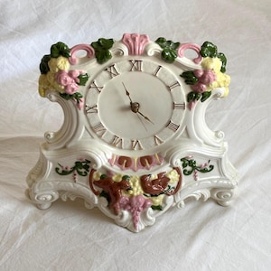 May include: A white ceramic mantel clock with a round face and Roman numerals. The clock is decorated with pink, yellow, and green floral accents. The clock measures approximately 8 inches wide and 9 inches tall.