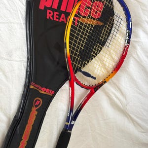 May include: A tennis racket with a black grip and a red, yellow, and blue frame, next to a black racket case. The racket has the word "Prince" in red on the frame and case. The case also has the words "Comfort Grip" and "Longbody".