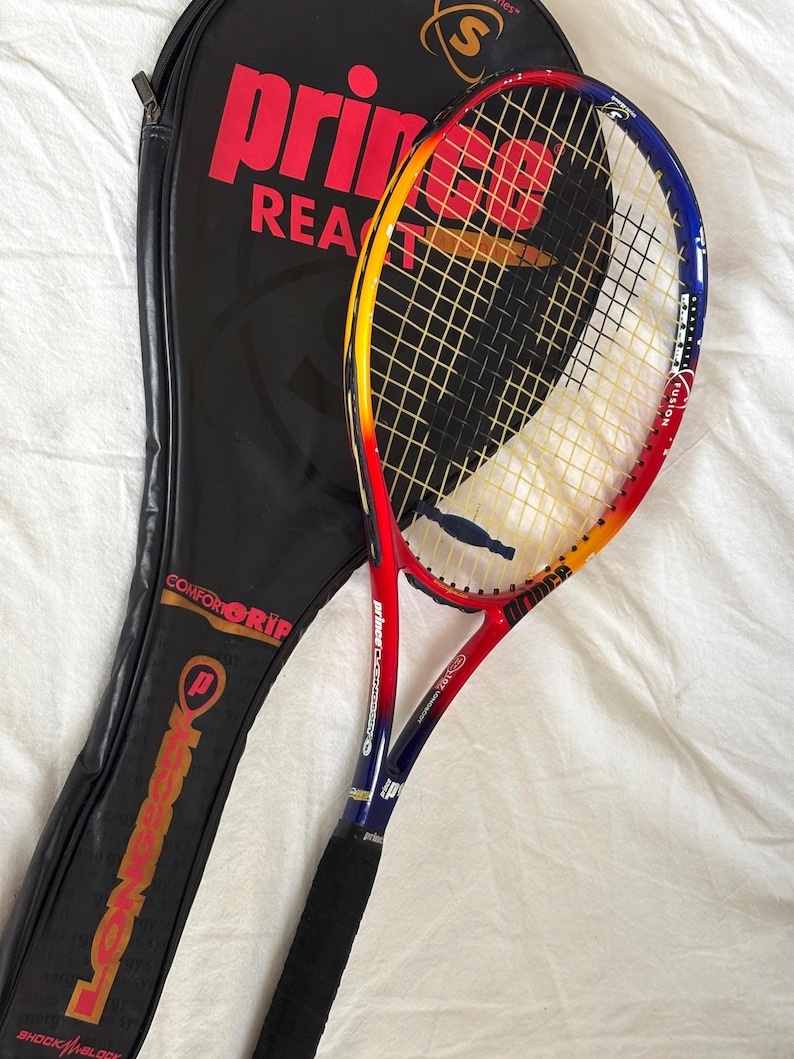 May include: A tennis racket with a black grip and a red, yellow, and blue frame, next to a black racket case. The racket has the word "Prince" in red on the frame and case. The case also has the words "Comfort Grip" and "Longbody".
