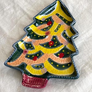 May include: A ceramic Christmas tree-shaped plate with a yellow and green design. The plate has a red base and is decorated with red and yellow dots. The edges are outlined in dark green and blue.