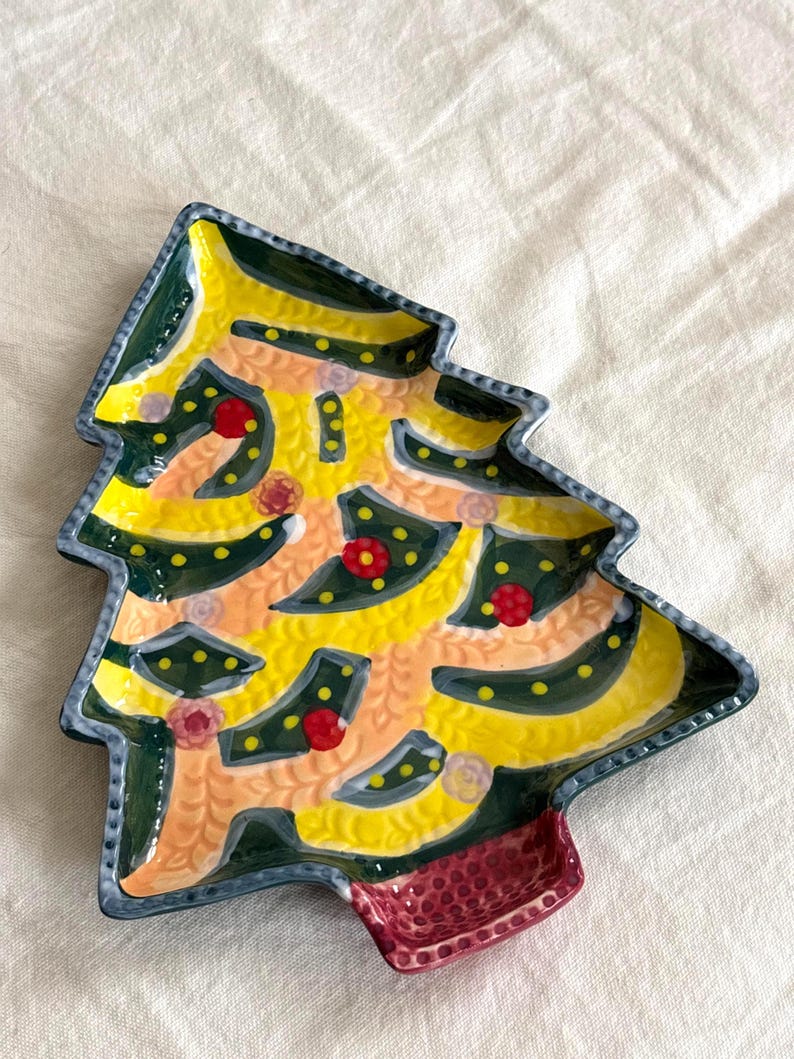 May include: A ceramic Christmas tree-shaped plate with a yellow, green, and red design. The plate has a textured surface and a small, rectangular section at the base. The plate is decorated with red and white floral accents.