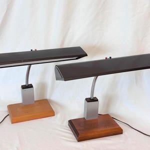 May include: Two desk lamps featuring long, dark brown rectangular shades, flexible silver metal necks, and rectangular wooden bases. Each lamp has a silver-toned vertical support column connecting the neck to the base and a black power cord.