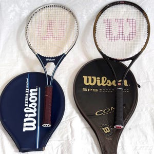 May include: Two Wilson tennis rackets with their covers. One racket has a silver frame and a navy blue handle, the other has a black frame. The covers are navy blue and black with the Wilson logo.