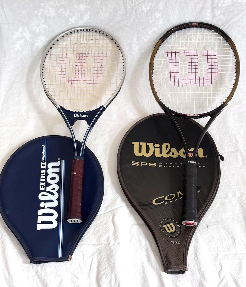 May include: Two Wilson tennis rackets with their covers. One racket has a silver frame and a navy blue handle, the other has a black frame. The covers are navy blue and black with the Wilson logo.