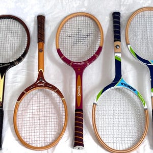May include: Five vintage tennis rackets with wooden frames and stringed nets. The rackets feature various handle designs and color schemes, including black, brown, red, and blue. Some rackets have brand names like "Spalding" and "Chemold."