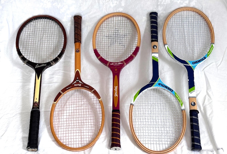 May include: Five vintage tennis rackets with wooden frames and stringed nets. The rackets feature various handle designs and color schemes, including black, brown, red, and blue. Some rackets have brand names like "Spalding" and "Chemold."