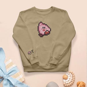 May include: A tan crew neck sweatshirt with a pixelated pink character design and a red mouth. The left sleeve has a small graphic and the letter "T". The sweatshirt is made of a soft material.