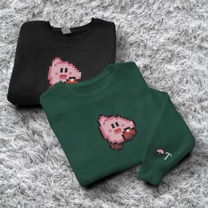 May include: Two sweatshirts, one black and one dark green, each with a pixelated pink character design. The green sweatshirt has a small embroidered design on the sleeve. Both are displayed on a textured, light grey surface.