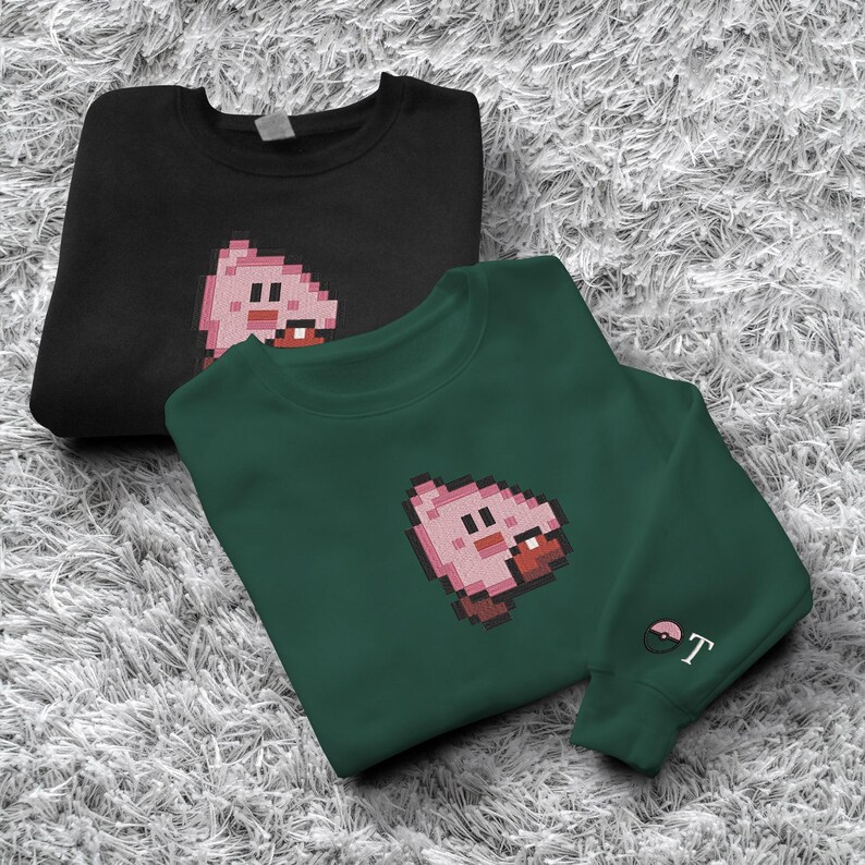 May include: Two sweatshirts, one black and one dark green, each with a pixelated pink character design. The green sweatshirt has a small embroidered design on the sleeve. Both are displayed on a textured, light grey surface.