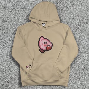 May include: A beige hoodie with a pixelated pink character design on the front. The hoodie has a hood, a front pocket, and a small logo on the left sleeve. The design is a cartoon-style image.