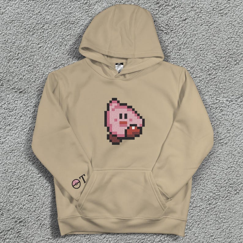 May include: A beige hoodie with a pixelated pink character design on the front. The hoodie has a hood, a front pocket, and a small logo on the left sleeve. The design is a cartoon-style image.