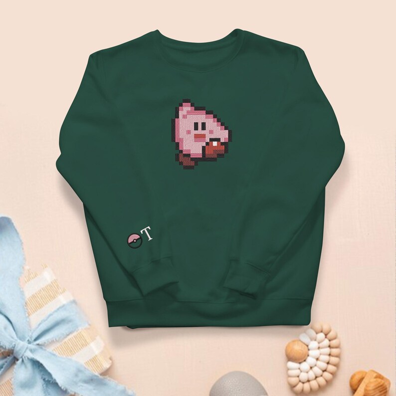 May include: A dark green sweatshirt with a pixelated pink character and red mouth on the front. The left sleeve has the white letter "T" next to a small, circular graphic. The sweatshirt is a comfortable, casual garment.