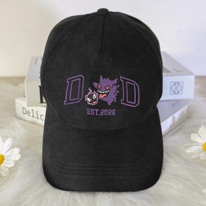 May include: Black baseball cap with a purple embroidered design featuring the letters "D&D" and two cartoon characters. The text "EST.2026" is below the characters. The cap has a curved brim and a button on top.