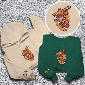 May include: A beige hooded sweatshirt and a green crew neck sweatshirt, each with an embroidered orange and yellow dragon design. The hoodie has a matching embroidered logo on the sleeve. A circular patch with the dragon design is also visible.
