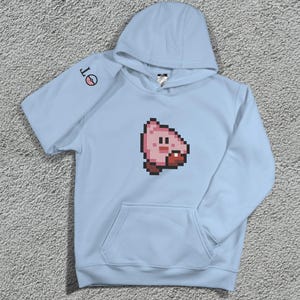 May include: A pale blue hoodie with a pixelated pink character on the front. The hoodie has a kangaroo pocket and a hood. The left sleeve features a small circular design with the letters "OT".