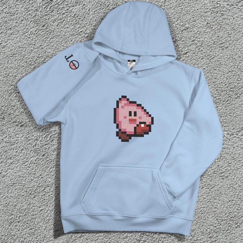 May include: A pale blue hoodie with a pixelated pink character on the front. The hoodie has a kangaroo pocket and a hood. The left sleeve features a small circular design with the letters "OT".