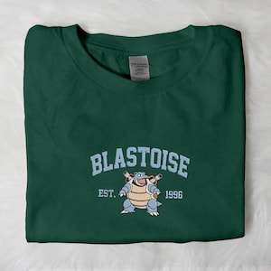 Può includere: T-shirt verde scuro con la scritta "BLASTOISE" in blu chiaro ad arco sopra un'illustrazione del personaggio. Sotto c'è la scritta "EST. 1996".